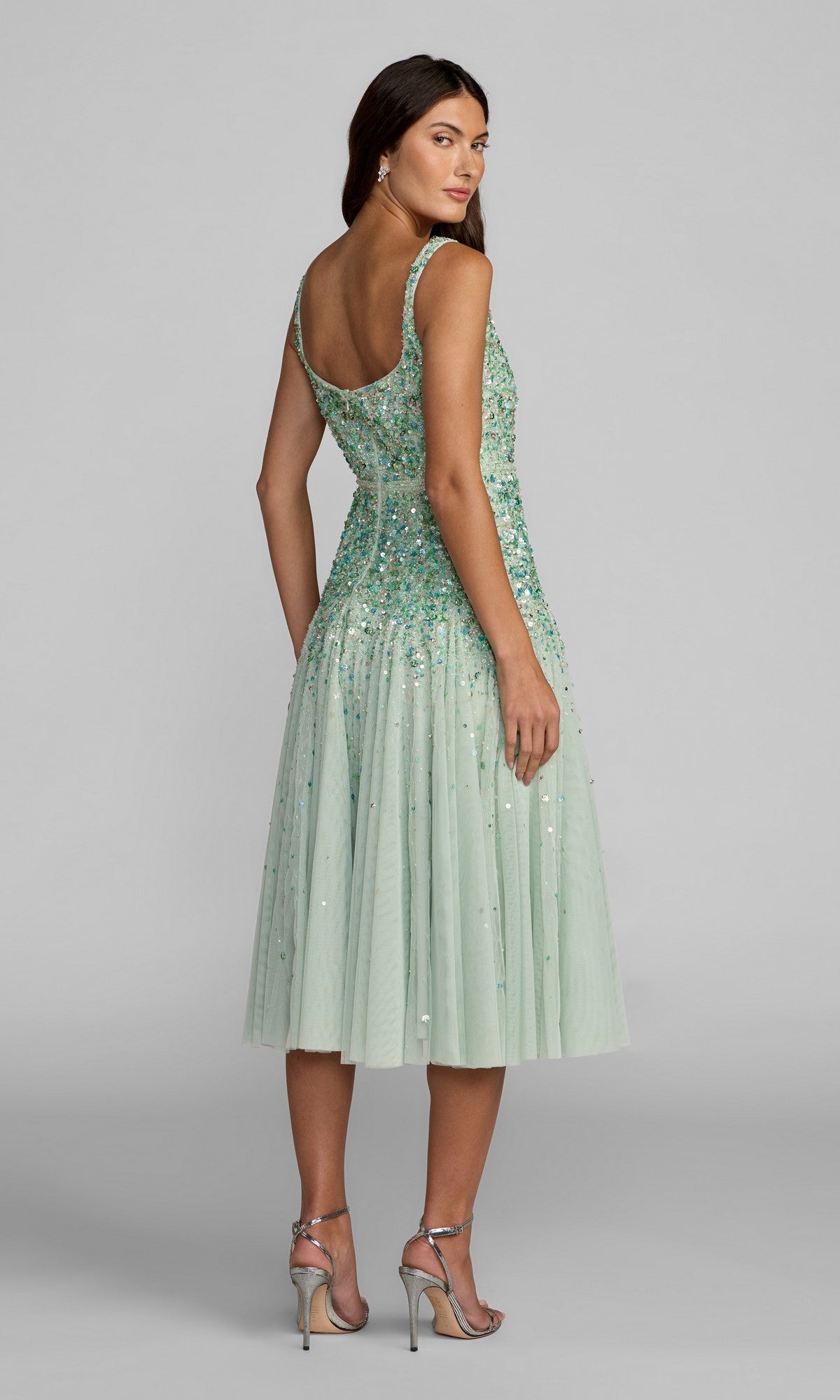 A woman stands facing away, slightly turning her head, in a Mac Duggal 94334 mint green sequin-embellished A-line midi wedding guest dress with thin straps and silver heels.